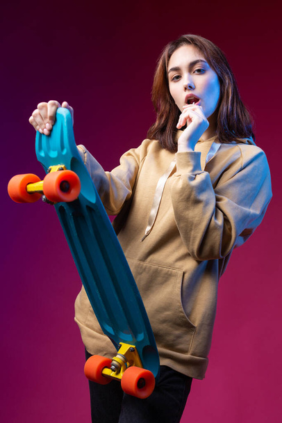 Joven chica hipster hermosa de moda vestida con una sudadera con capucha sostiene un monopatín en sus manos sobre un fondo de estudio púrpura. Come una piruleta.. - Foto, Imagen