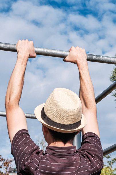 Back view of a young man in a hat pull-up on the bar. Hipster or nerd exercising outdoors. - Photo, Image