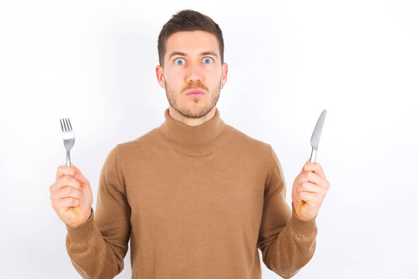 hungry young caucasian man wearing turtleneck over white background holding in hand fork knife want tasty yummy pizza pie - Photo, Image