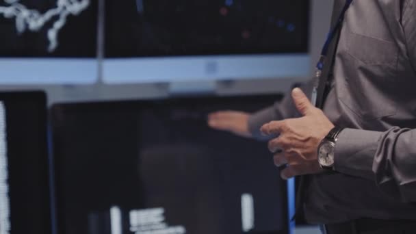 Basso angolo di taglio specialista di cyber sicurezza maschile indossa camicia grigia, in piedi in ufficio moderno, spiegando compiti ai colleghi irriconoscibili, indicando monitor del computer - Filmati, video