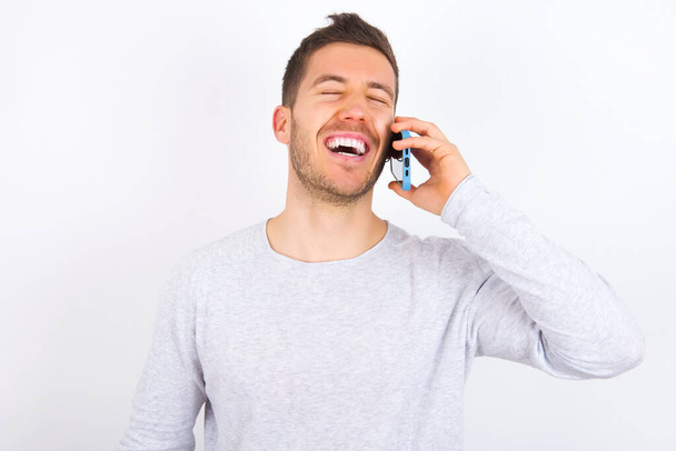 Overemotive happy young caucasian man wearing casual clothes over white background laughs out positively hears funny story from friend during telephone conversation - Foto, imagen