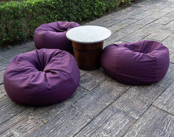 Tavolino rotondo vuoto in marmo con tre sacchetti di fagioli viola a terra vicino al cespuglio verde, decorazione del giardino all'aperto. - Foto, immagini