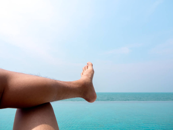Primer plano del pie del hombre con las piernas cruzadas en el agua azul en la piscina tropical y vista al mar. Vacaciones, vacaciones fondo de verano con espacio de copia. - Foto, imagen