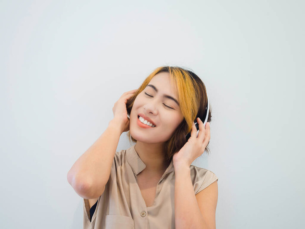Портрет щасливої азіатки з коротким волоссям у бежевій сорочці полюбляє слухати музику або пісню по радіо з білим навушником на білому тлі, закриваючи очі.. - Фото, зображення