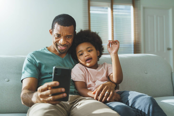 Smiling African American father and his cute boy using smartphone at home, African family doing video call on mobile phone, Black Dad and son sitting on sofa taking selfies. - Фото, зображення