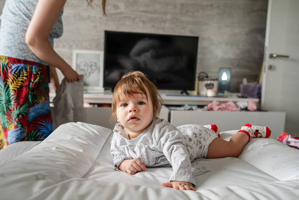 Kaukasisches Baby liegt zu Hause auf dem Bett im hellen Zimmer auf dem Bauch nach unten, während ihre Mutter hinter ihr steht und nach Kleidungsstücken sucht, um die Kindheit und das Familienkonzept echter Menschen zu verändern. - Foto, Bild