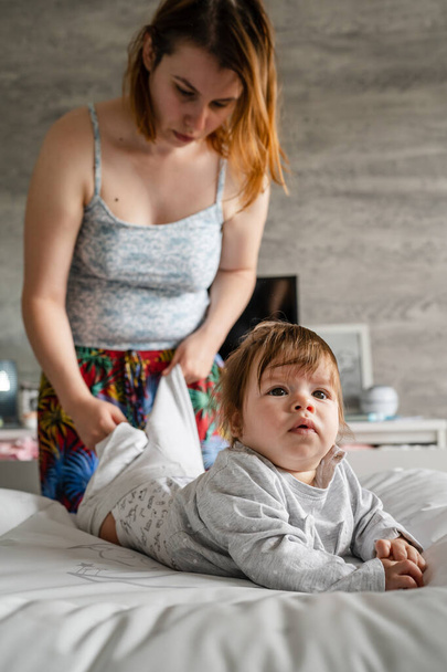 Caucasian baby lying on the bed at home in the bright room on the belly down while her mother is standing behind looking for clothes to change real people childhood and family concept - Foto, imagen