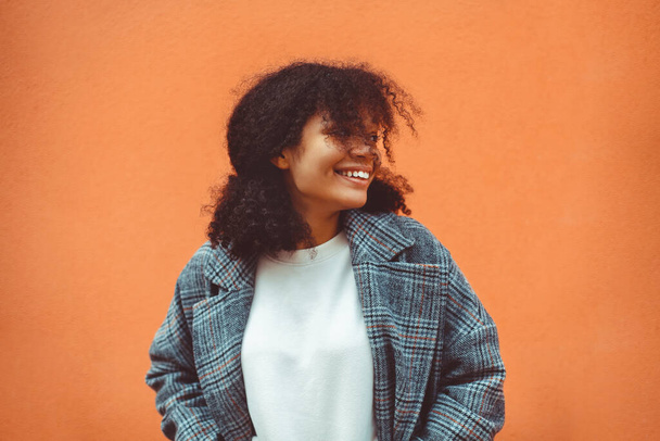 Hayatın tadını çıkarıyorum. Güzel mutlu Afrikalı etnik kız koyu kıvırcık saçlı, şık montlu, turuncu duvarın arkasında duran, geniş, parlak bir gülümsemeyle başka tarafa bakan. Mutluluk kavramı - Fotoğraf, Görsel