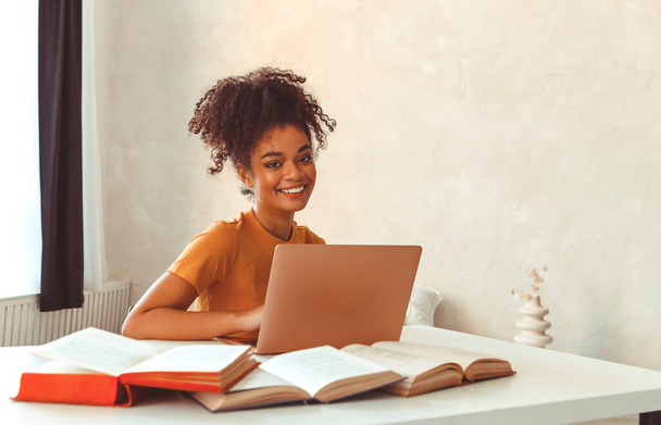 Весела молода афроамериканська студентка сидить за столом у оточенні підручників, перед ноутбуком навчається старанно готуватися до іспитів онлайн, дивлячись в камеру з задоволеною посмішкою - Фото, зображення