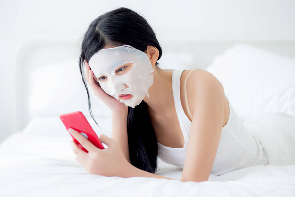 Schöne junge asiatische Frau mit Laken Gesichtsmaske liegend Smartphone auf dem Bett im Schlafzimmer, Schönheit Mädchen Anwendung Gesichtsmaske für Hautpflege von Falten zu Hause, Hautpflege, Gesundheit und Wohlbefinden. - Foto, Bild