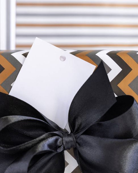 Present with blank square gift tag and black ribbon bow close up. Gift boxes wrapped in Striped and chevron geometric paper. Christmas, New Year, Birthday, Anniversary label mockup - Zdjęcie, obraz