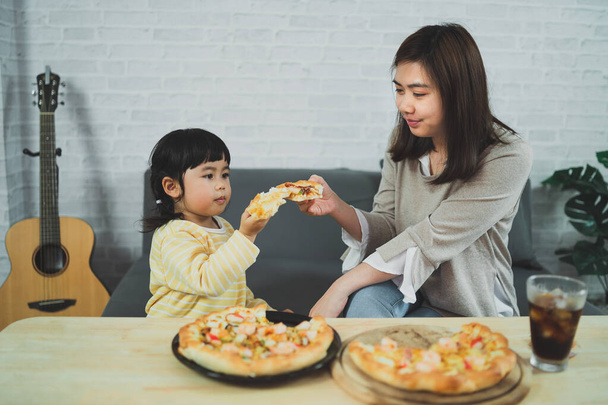 アジアの母親と子供たちは自宅で活動しています。母親は子供たちのためにピザを食べます。子供たちはイタリアの自家製ピザを食べて試飲しています。かわいい子供たちは居心地の良い家でおいしい料理を楽しんでいます。ウイルスの流行の間に、 Covid-19 - 写真・画像