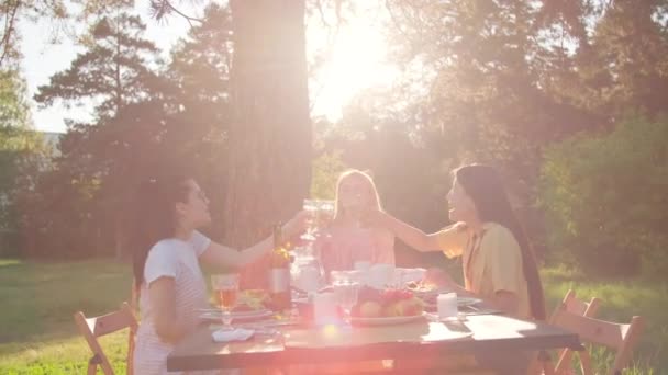 Группа из трех молодых женщин в стильной повседневной одежде обедают вместе на открытом воздухе в парке, звонят в стаканы, празднуя что-то - Кадры, видео