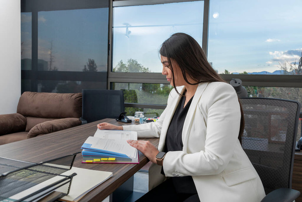 Mulher de negócios lendo um livro em sua mesa de escritório com janelas no fundo. Jovem latina lê enquanto trabalha em seu escritório. - Foto, Imagem