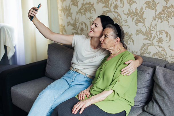 Grandmother and granddaughter look at the smartphone while sitting on the couch. Taking a selfie on the phone - Фото, изображение