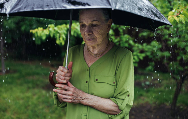 An old woman stands on the street under the rain with a black umbrella - Foto, imagen