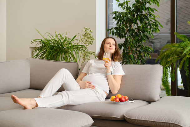 Cute pregnant woman having rest sitting on sofa at home with a glass of fresh orange juice in hand - Foto, imagen