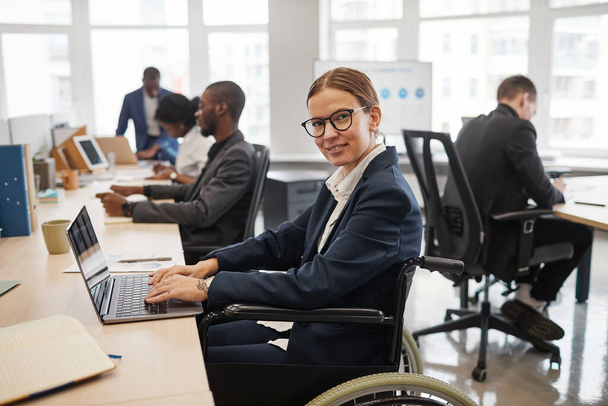 Retrato vertical de vista lateral de una joven mujer de negocios que usa silla de ruedas mientras trabaja con una computadora portátil en la oficina - Foto, imagen