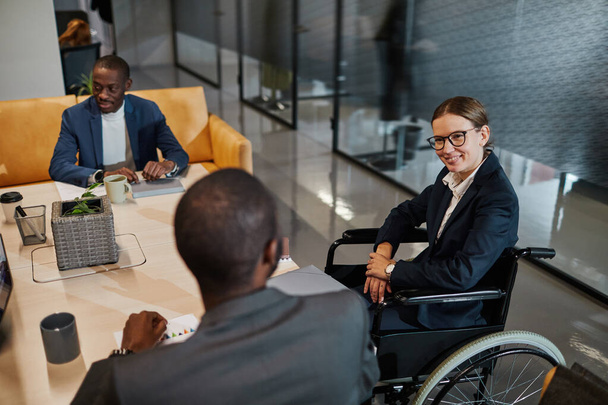 Портрет успешной предпринимательницы в инвалидной коляске на встрече и беседе с группой коллег в современном офисном пространстве - Фото, изображение