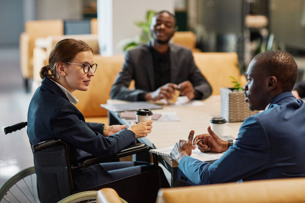 Портрет успішної бізнес-леді, яка використовує інвалідне крісло на зустрічі і розмовляє з групою колег в сучасному офісному просторі
 - Фото, зображення