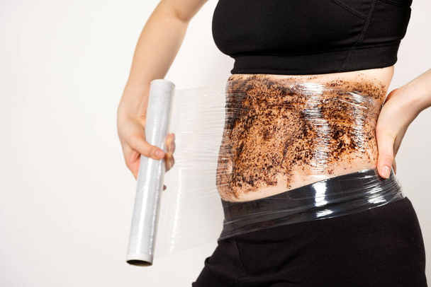 Μια γυναίκα κάνει ένα περιτύλιγμα κοιλιάς με ένα αντικυτταρικό τρίψιμο σώματος καφέ. Η έννοια της απώλειας βάρους και φροντίδα του δέρματος - Φωτογραφία, εικόνα