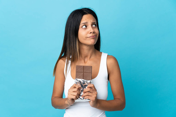 Valkoihoinen tyttö eristetty sinisellä pohjalla ottaen suklaa tabletti ja joilla on epäilyksiä - Valokuva, kuva
