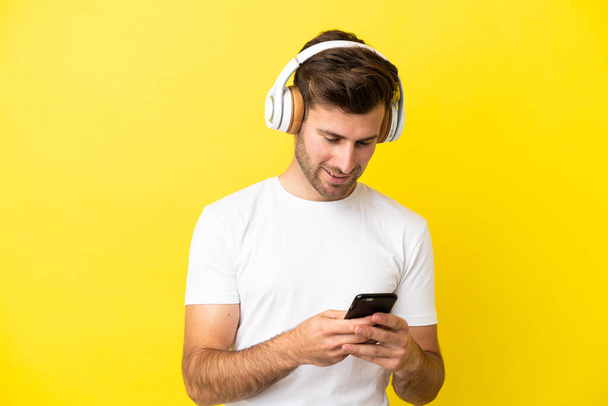 Young caucasian handsome man isolated on yellow background listening music and looking to mobile - Фото, изображение