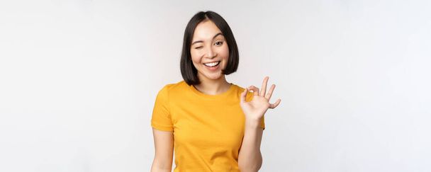 Hermosa mujer joven mostrando signo bien, sonriendo complacido, recomendando smth, aprobar, como producto, de pie en camiseta amarilla sobre fondo blanco - Foto, imagen