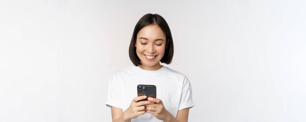Портрет усміхненої азіатки, що використовує мобільний телефон, балачки, смс-повідомлення, стоїть у сорочці на білому тлі
 - Фото, зображення
