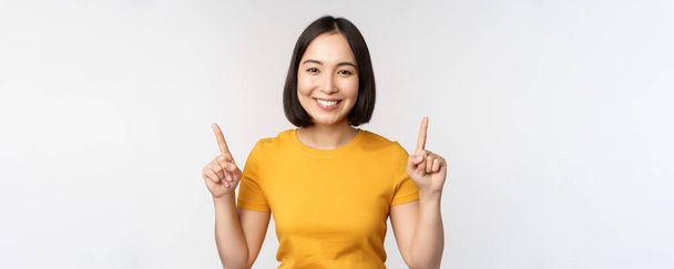 Portrait de belle fille japonaise souriante, pointant les doigts vers le haut, montrant la publicité, debout en tshirt jaune sur fond blanc - Photo, image