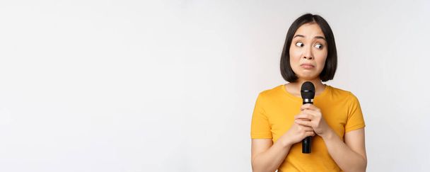 Modest asian girl holding microphone, scared talking in public, standing against white background - Photo, image
