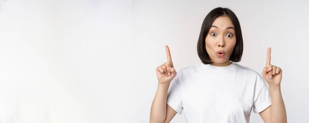 Portrait of enthusiastic young woman, asian girl smiling pointing fingers up, showing advertisement upwards, standing over white background - Foto, imagen