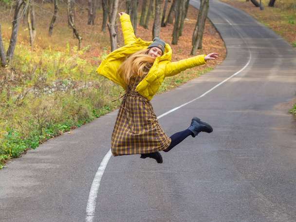 Modern kıyafetli gülen kadın sonbahar parkında dar bir yolda zıplıyor. - Fotoğraf, Görsel