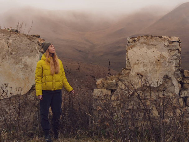 Una mujer está de pie entre la hierba seca de otoño cerca de las paredes de una casa en ruinas contra el fondo de las colinas con niebla que desciende sobre ellos. - Foto, Imagen