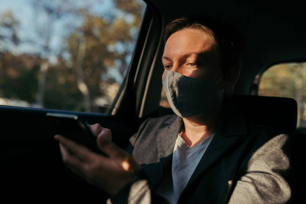 Businesswoman typing text message in car while wearing protective face mask for Covid-19 pandemic, selective focus - Photo, image