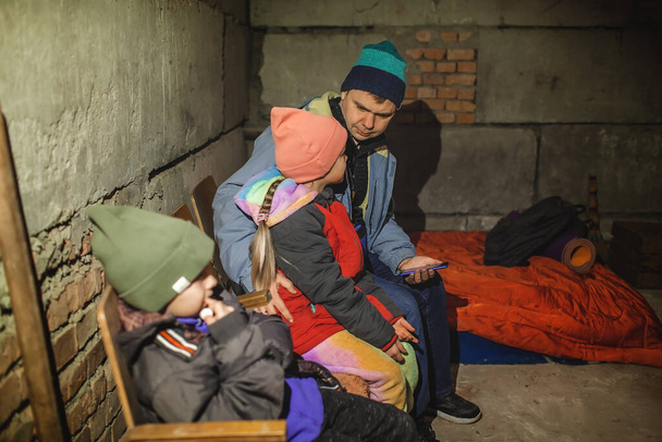子供とウクライナの父は爆弾の避難所に座って、ロシアの侵略者に関する最新のブレーキニュースを読む - 写真・画像