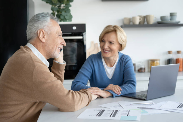 Portrait of retired couple sitting in modern kitchen. Husband talking to phone. Solving financial issues. Utility bills document. Planning family budget. Calculating expenses - Photo, image