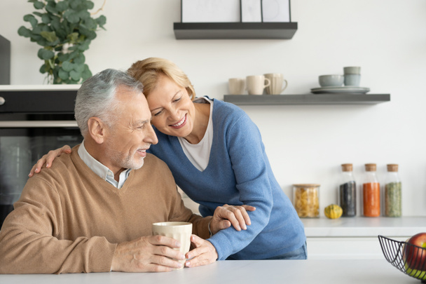 Χαμογελώντας και τρυφερή συνταξιούχος σύζυγος αγκαλιάζει τον άντρα της. Πίνω πρωινό καφέ ή τσάι. Σπιτική άνεση. Έννοια παντοτινής αγάπης. Νιώθοντας ευτυχισμένοι μαζί - Φωτογραφία, εικόνα
