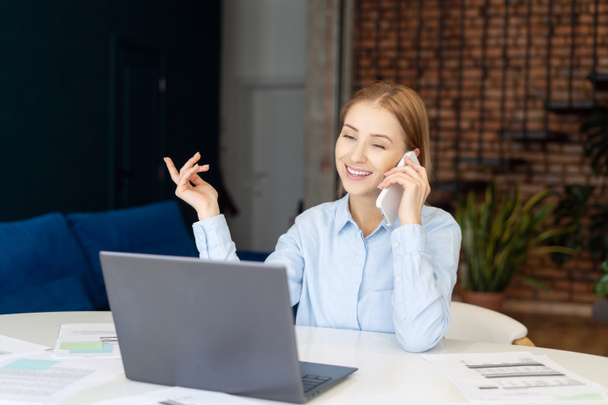 Portrait of smiling woman talking to phone with customer. Home office concept. Comfortable workplace with laptop during pandemic. Communication with business partners using gadgets. Remote job - Photo, image