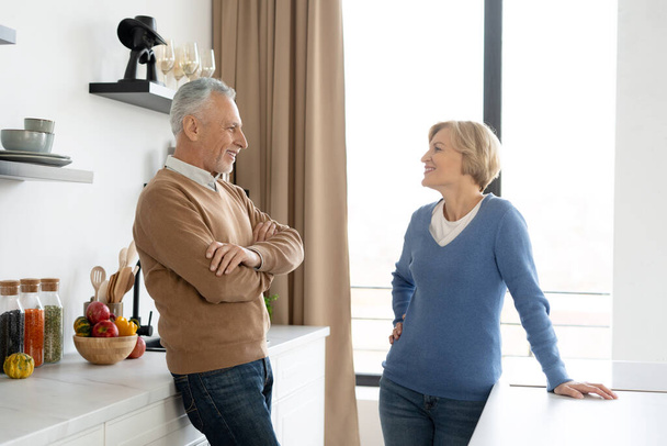 Vue latérale du couple souriant d'âge moyen regardant parler dans la cuisine moderne confortable. Bonne famille. Concept d'amour éternel. Passer le week-end ensemble - Photo, image
