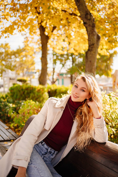 a beautiful blonde in a coat and jeans on the bench in an autumn park.  - Photo, image