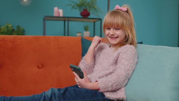 Boldog kis iskolás lány használja mobiltelefon csinál győztes gesztus ünnepelni egyedül otthon kanapé - Fotó, kép