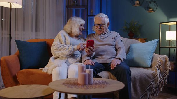 Старша пара дідусь бабуся робить онлайн покупки на мобільному телефоні, сидячи на домашньому дивані
 - Фото, зображення