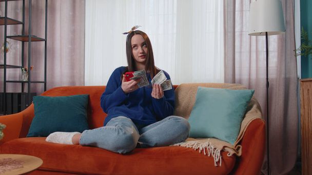 Улыбающаяся счастливая молодая женщина с деньгами и мобильным телефоном рассчитывает домашние счета дома - Фото, изображение