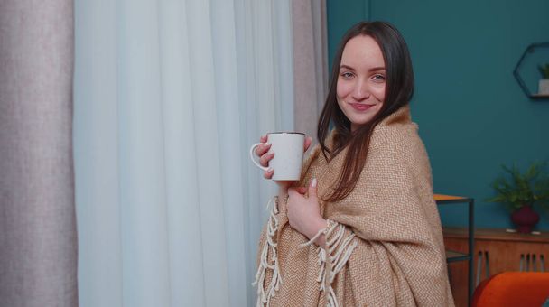 jong volwassen brunette vrouw in warm wit trui drinken warm drankje mok ontspannen thuis in de buurt venster - Foto, afbeelding