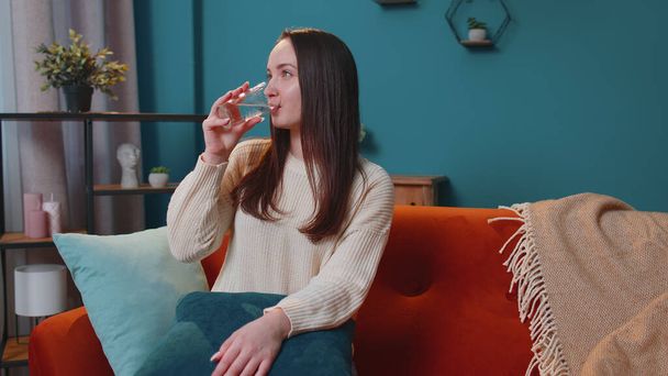 Sede uma jovem mulher sentada em casa segurando vidro de aqua natural fazer goles beber água parada - Foto, Imagem