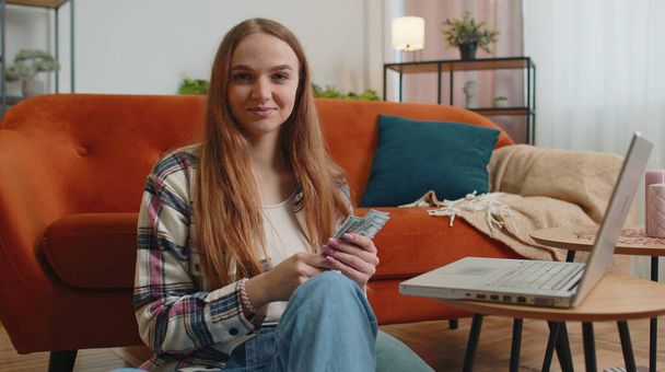 Улыбающаяся счастливая молодая женщина, подсчитывающая деньги и использующая ноутбук, рассчитывает домашние счета дома - Фото, изображение