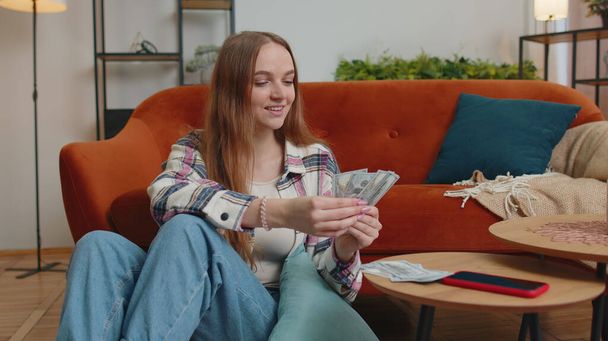 Улыбающаяся счастливая молодая женщина считает деньги, рассчитывает домашние счета дома удовлетворенные доходом - Фото, изображение