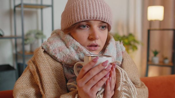 Young sick woman wear hat wrapped in plaid sit alone shivering from cold on sofa drinking hot tea - Foto, Imagem
