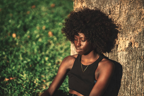 A portrait of a cute pensive young black lady with fluffy curly hair, leaning against the tree while sitting on the lawn of a park on a warm sunny day, a copy space place on the left for an ad message - Foto, immagini